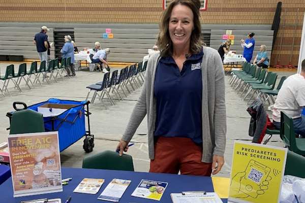 A woman standing inside next to a table with A1C materials, and smiling at the camera.
