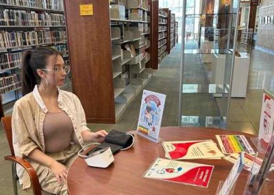 A young woman sits at a wooden table inside a library, preparing to use a self-service blood pressure monitor. The table is stocked with health brochures and instructional signs, with rows of bookshelves visible in the background.