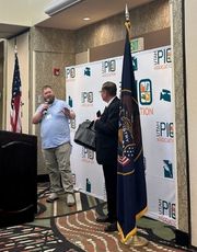 Two men speaking at a professional presentation next to a podium and a large backdrop banner displaying event logos, with the American and state flags standing nearby.